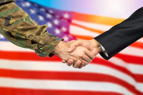 American soldier in uniform and civil man in suit shaking hands with adequate national flag on background